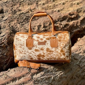 May include: A brown and white cowhide duffel bag with leather handles and accents. The bag has a detachable shoulder strap and a small rectangular tag. The bag is sitting on a rocky surface.