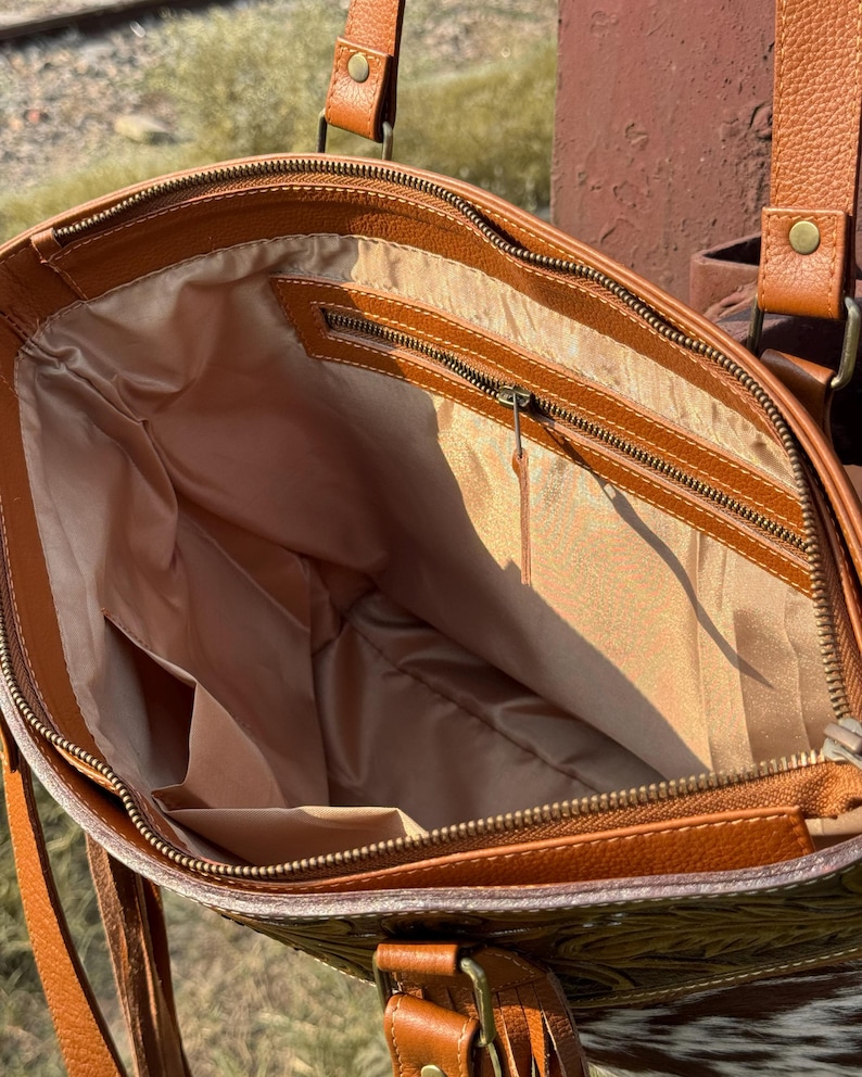 Pode incluir: Vista aberta de uma bolsa tote de couro marrom com fecho de z&iacute;per. A bolsa tem um interior marrom claro com v&aacute;rios bolsos. O exterior apresenta um padr&atilde;o em relevo e al&ccedil;as de couro. A bolsa tem um tema western.