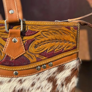 Pode incluir: Close-up de uma bolsa de couro marrom com um design detalhado de penas. A bolsa apresenta uma parte inferior de couro de vaca com uma mistura de p&ecirc;lo branco e castanho. A bolsa tem uma al&ccedil;a de couro castanho e ferragens prateadas.