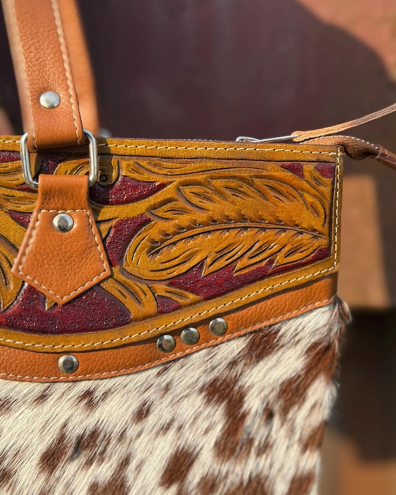 Pode incluir: Close-up de uma bolsa de couro marrom com um design detalhado de penas. A bolsa apresenta uma parte inferior de couro de vaca com uma mistura de p&ecirc;lo branco e castanho. A bolsa tem uma al&ccedil;a de couro castanho e ferragens prateadas.