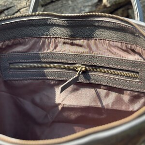 K&ouml;nnte beinhalten: Nahaufnahme des Innenraums einer braunen Leder-Tragetasche. Die Tasche hat eine Rei&szlig;verschlusstasche mit einem Messingrei&szlig;verschluss und einem braunen Leder-Zipper. Das Innenfutter ist hellbraun. Die Tasche hat eine strukturierte Au&szlig;enseite und einen grauen Griff.