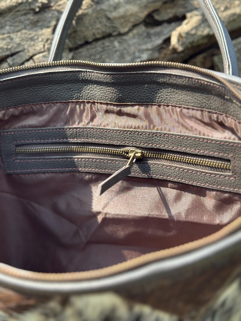 K&ouml;nnte beinhalten: Nahaufnahme des Innenraums einer braunen Leder-Tragetasche. Die Tasche hat eine Rei&szlig;verschlusstasche mit einem Messingrei&szlig;verschluss und einem braunen Leder-Zipper. Das Innenfutter ist hellbraun. Die Tasche hat eine strukturierte Au&szlig;enseite und einen grauen Griff.