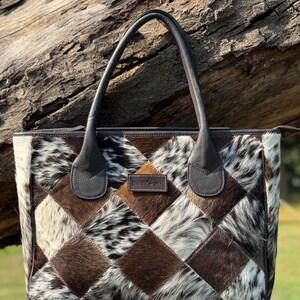 K&ouml;nnte beinhalten: Patchwork-Tragetasche mit einem geometrischen Muster aus braunen, wei&szlig;en und schwarzen Kuhfellquadraten. Die Tasche hat dunkelbraune Ledergriffe und -verzierungen. Die Tasche ist rechteckig und hat einen Rei&szlig;verschluss.