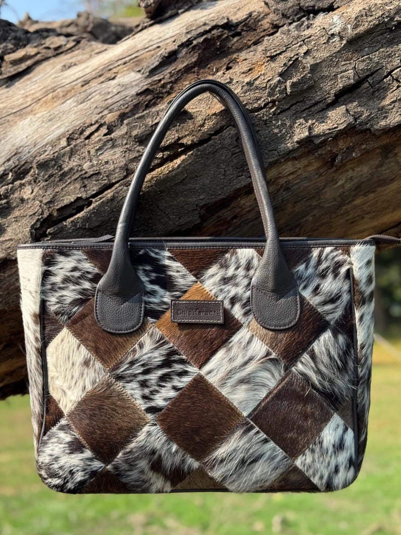 K&ouml;nnte beinhalten: Patchwork-Tragetasche mit einem geometrischen Muster aus braunen, wei&szlig;en und schwarzen Kuhfellquadraten. Die Tasche hat dunkelbraune Ledergriffe und -verzierungen. Die Tasche ist rechteckig und hat einen Rei&szlig;verschluss.