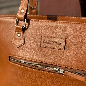 Pode incluir: Close-up de uma mala tote de couro castanho com uma superf&iacute;cie texturizada. A mala apresenta um fecho de correr prateado e uma pequena etiqueta retangular com o texto "Chic Harbor". A mala tem uma pega e uma al&ccedil;a.