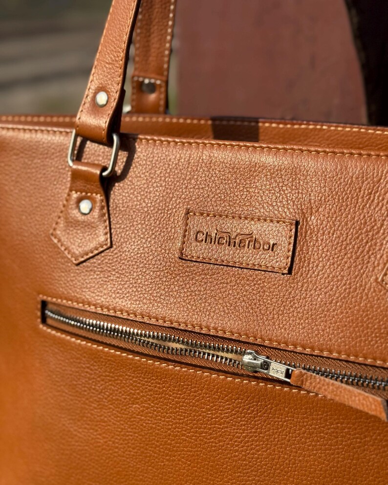 Pode incluir: Close-up de uma mala tote de couro castanho com uma superf&iacute;cie texturizada. A mala apresenta um fecho de correr prateado e uma pequena etiqueta retangular com o texto "Chic Harbor". A mala tem uma pega e uma al&ccedil;a.