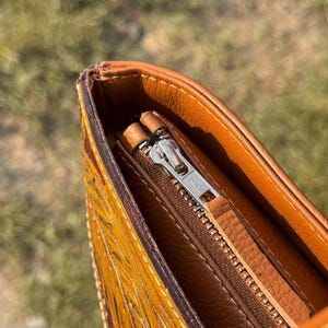 Pode incluir: Close-up de uma bolsa de couro marrom com z&iacute;per prateado. A bolsa tem um design detalhado e texturizado em tons de amarelo e vermelho. O forro interno &eacute; de um tom marrom mais claro.