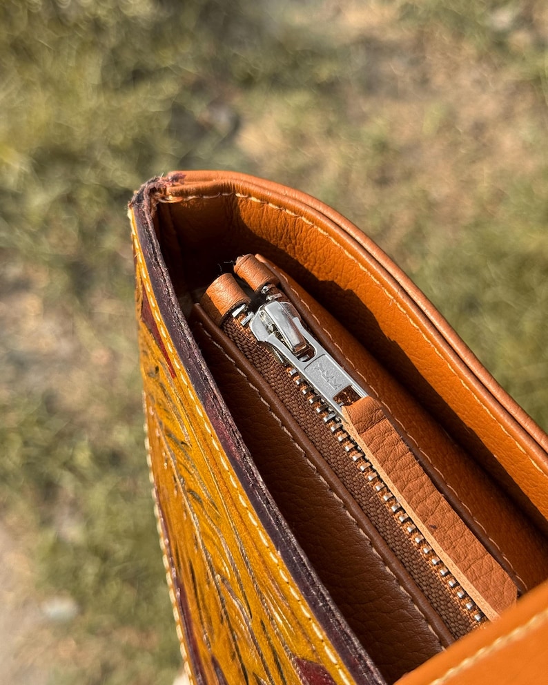 Pode incluir: Close-up de uma bolsa de couro marrom com z&iacute;per prateado. A bolsa tem um design detalhado e texturizado em tons de amarelo e vermelho. O forro interno &eacute; de um tom marrom mais claro.