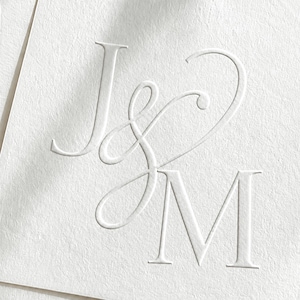 May include: A white card featuring a blind embossed monogram of the letters J and M. The letters are pressed into the textured paper surface, creating a subtle three-dimensional effect with shadows that define the elegant serif and script typography.