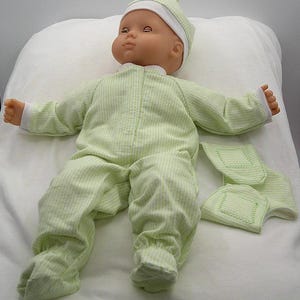 May include: A baby doll with a light skin tone, wearing a light green and white gingham sleeper with long sleeves and footed bottoms. The doll also wears a matching light green and white gingham hat with a small knot on top. A separate matching fabric piece is included.