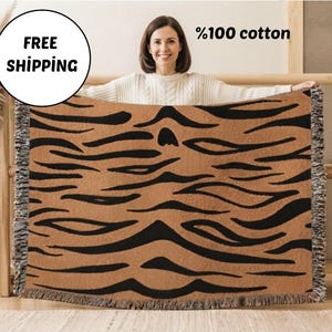 May include: A rectangular woven throw blanket featuring a repeating black tiger stripe pattern on a tan background. The blanket is made of 100% cotton and has decorative fringe along the left and right vertical edges of the fabric.
