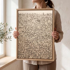 May include: A framed art print with a detailed black and white illustration of a crowd of people. The artwork has a beige background and is in a natural wood frame. The print is held by a person.