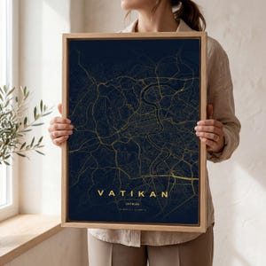 May include: A framed map print of Vatican City. The map shows gold lines on a dark blue background. The word "VATIKAN" is printed in gold at the bottom, with the city's coordinates below. The frame is made of light-coloured wood.