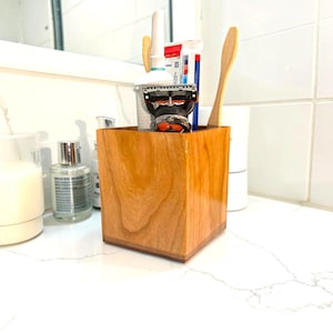 May include: A square wooden bathroom organizer with a polished finish and visible wood grain. The container holds a metal razor, a tube of toothpaste, and a bamboo toothbrush, providing a storage solution for various personal care items.