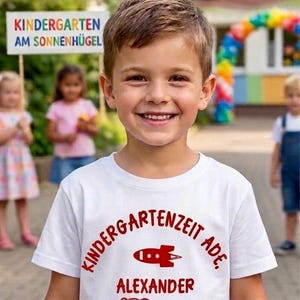Könnte beinhalten: Ein lächelnder Junge trägt ein weißes T-Shirt mit rotem Text und Grafiken. Auf dem Shirt steht "Kindergartenzeit Ade", "Alexander" und "ABC 123". Im Hintergrund steht ein Schild mit der Aufschrift "Kindergarten am Sonnenhügel".
