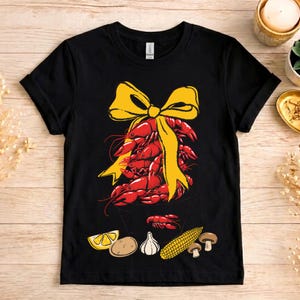 Peut inclure: T-shirt noir avec un motif de langoustes rouges attachées avec un nœud jaune. En dessous, des illustrations de tranches de citron, d'une pomme de terre, d'ail, de maïs et de champignons. Le t-shirt est à col rond.