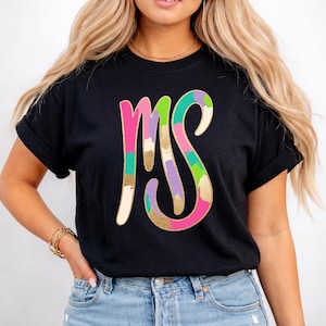 Peut inclure: T-shirt noir avec les lettres "MS" dans un design coloré au pinceau. Les lettres sont roses, vertes, violettes et dorées, avec un effet pailleté doré. Le t-shirt est fait d'une matière douce.