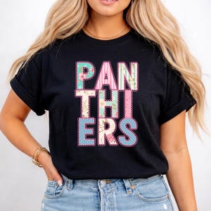 Peut inclure: T-shirt noir avec le mot "PANTHERS" dans un design coloré, floral et à rayures. Les lettres sont soulignées en rose et ont un bord festonné. Le t-shirt est fait d'une matière douce.