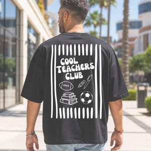Könnte beinhalten: Ein schwarzes Kurzarm-T-Shirt mit einer weißen Grafik auf der Rückseite. Das Design zeigt vertikale Streifen, den Text "COOL TEACHERS CLUB" sowie Illustrationen eines Footballs, eines Textmarkers, eines Stifts, eines Bücherstapels und eines Fußballs.