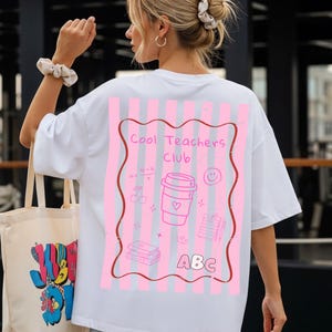Könnte beinhalten: Ein weißes T-Shirt mit einer Grafik auf der Rückseite, bestehend aus rosa vertikalen Streifen und einem gewellten braunen Rand. Das Design enthält den Text "Cool Teachers Club" und "ABC" sowie Abbildungen eines Kaffeebechers, Smileys, Büchern und Kirschen.