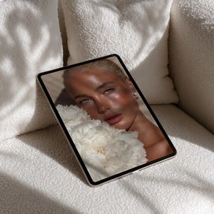May include: A digital tablet displaying a close-up of a person with blonde hair and blue eyes, holding white flowers. The tablet rests on a white, textured sofa, with sunlight casting shadows.