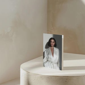 May include: A greeting card featuring a woman in a white satin robe, standing on a white surface. The card is propped up on a textured, light-colored concrete pedestal. The background is a neutral beige.