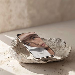 May include: A smartphone with a gold-colored frame displaying a portrait of a person with blue eyes and freckles. The phone rests on a rough, light-colored stone, with shadows cast across the surface.