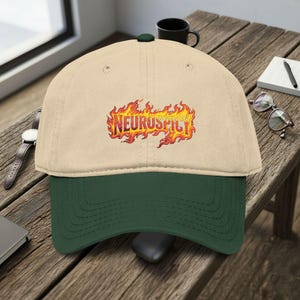 May include: A beige and green baseball cap with the word "NEUROSPICY" in a fiery, orange and yellow font. The cap has a curved brim and a dark green underside. The cap is sitting on a wooden surface.