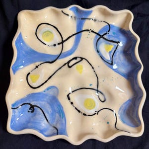 May include: A square ceramic dish with a wavy, scalloped edge and a cream-coloured base. The surface features hand-painted blue watercolour-style washes, small yellow circular accents, and thin, continuous black lines that loop across the plate.