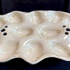 May include: An oval ceramic egg serving tray with a glossy off-white glaze and mottled beige speckles. The tray features twelve recessed egg-shaped indentations arranged in two rows, with three small circular holes positioned on each side.
