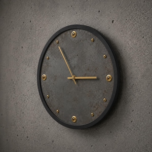 Könnte beinhalten: Eine runde Wanduhr mit einem dunkelgrauen, texturierten Beton-Zifferblatt in einem dünnen schwarzen Rahmen. Die Uhr hat goldfarbene Zeiger und goldfarbene Metallschrauben, die als Stundenmarkierungen am Rand des Zifferblatts dienen.