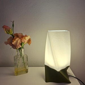 Może przedstawiać: Nowoczesna lampa stołowa z białym kloszem i ciemnozieloną podstawą. Lampa jest włączona, emitując miękką, ciepłą poświatę. Obok lampy znajduje się przezroczysta szklana butelka z kwiatami w kolorze brzoskwini.