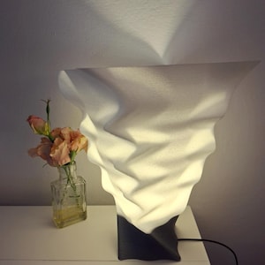 Może przedstawiać: Nowoczesna biała lampa stołowa o unikalnym, teksturowanym designie. Lampa ma ciemnoszarą podstawę i biały, falisty klosz. W pobliżu stoi mały szklany wazon z kwiatami w kolorze brzoskwini.