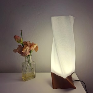 Może przedstawiać: Nowoczesna lampa stołowa z białym, teksturowanym abażurem i brązową podstawą. Lampa jest podświetlona, emitując ciepłą poświatę. W pobliżu stoi mała szklana butelka z brzoskwiniowymi kwiatami.