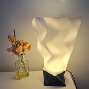 Może przedstawiać: Nowoczesna biała lampa stołowa o unikalnym, teksturowanym designie. Lampa ma czarną podstawę i miękkie, świecące światło. Obok lampy stoi mała szklana butelka z pomarańczowymi i różowymi kwiatami.