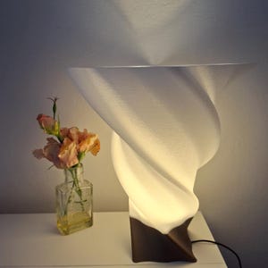 Może przedstawiać: Nowoczesna biała lampa stołowa o wirującym wzorze i brązowej podstawie. Lampa jest podświetlona, emitując ciepłą poświatę. W pobliżu stoi mały szklany wazon z kwiatami w kolorze brzoskwini.