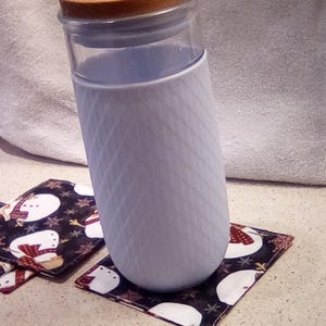 Könnte beinhalten: Ein durchsichtiger Glasbecher mit hellblauem Silikonmantel und Holzdeckel. Der Becher steht auf einem quadratischen Untersetzer mit schwarzem Hintergrund und Schneemannmuster. Ein klarer Strohhalm ist in den Deckel eingesetzt.
