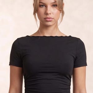Peut inclure: Un haut noir à manches courtes doté d'une encolure bateau avec une finition subtile à bords ondulés. Le vêtement présente des détails froncés sur les côtés créant une texture plissée le long du torse, finissant par un ourlet asymétrique.