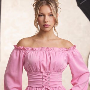 Può includere: Un top rosa con spalle scoperte, maniche lunghe e vita stretta. Il top presenta uno scollo a balze e un dettaglio di allacciatura in stile corsetto al centro. Il tessuto ha una leggera lucentezza e lo stile generale è femminile ed elegante.
