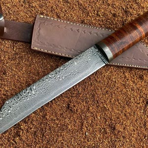Puede incluir: Un cuchillo de acero de Damasco con una hoja estampada y un mango de madera. El cuchillo descansa sobre una funda de cuero marrón con una correa de cuero a juego. El mango es de color marrón oscuro.