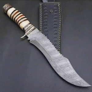 Puede incluir: Un cuchillo de caza grande y ornamentado con una hoja estampada y un mango decorativo con detalles de hueso y madera. El cuchillo va acompañado de una funda de cuero negro con costuras decorativas. La hoja tiene un borde curvo y un lomo dentado.