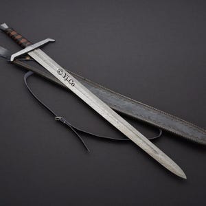 Puede incluir: Una espada larga y plateada con un mango envuelto en cuero marrón y una guarda cruzada. La espada está en una funda de cuero negro con una correa. El texto "© Yj.Co" está impreso en la hoja. El fondo es negro.