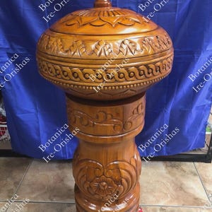 May include: A handcrafted, wooden pedestal with a matching lidded bowl. The bowl and pedestal are intricately carved with floral and geometric patterns. The wood has a warm, brown tone, and the piece is displayed against a blue backdrop.