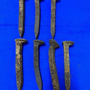 May include: Seven weathered, rusty railroad spikes with rectangular heads and tapered points are arranged against a blue background. The spikes show signs of age and use, with a dark brown and gray color.