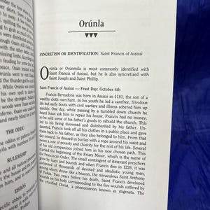 May include: Open book with text about Or&uacute;nla and Saint Francis of Assisi. The text is in black font on white pages. The book is open on a blue surface.