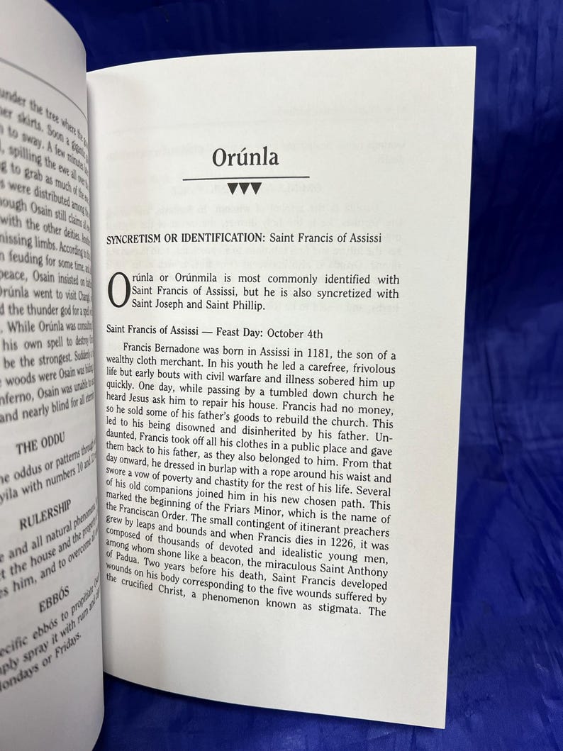 May include: Open book with text about Or&uacute;nla and Saint Francis of Assisi. The text is in black font on white pages. The book is open on a blue surface.