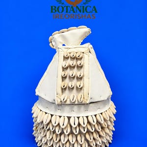 May include: A white leather ceremonial headpiece adorned with numerous cream-colored cowrie shells arranged in rows and a vertical panel. The text BOTANICA IREORISHAS appears above the object. The headpiece features a structured, layered design.