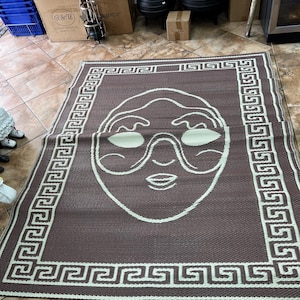 May include: Brown and white rectangular mat with a face design. The mat features a border with a Greek key pattern and a central illustration of a face with glasses. The mat is made of woven material.