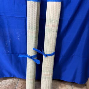 May include: Two rolled-up woven mats with blue trim and blue ribbon ties. The mats are beige with green and red accents. The background is a solid blue fabric.