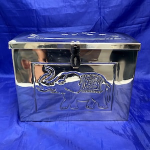 May include: A shiny silver metal trunk with a rectangular shape. The front of the trunk features a raised relief of an elephant. The trunk has a hinged lid and a latch closure.
